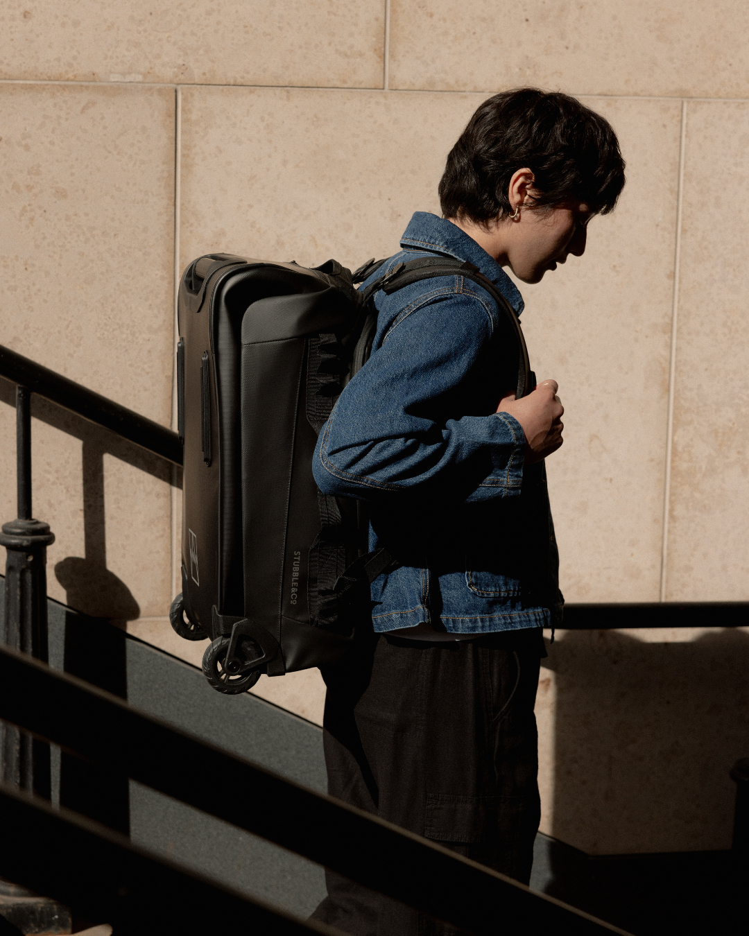 Woman in a denim jacket wearing the Wheeled Duffel in All Black as a backpack.