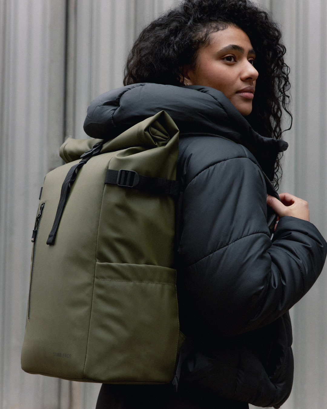 A woman wearing a black puffer jacket and a Roll Top Backpack in Urban Green.