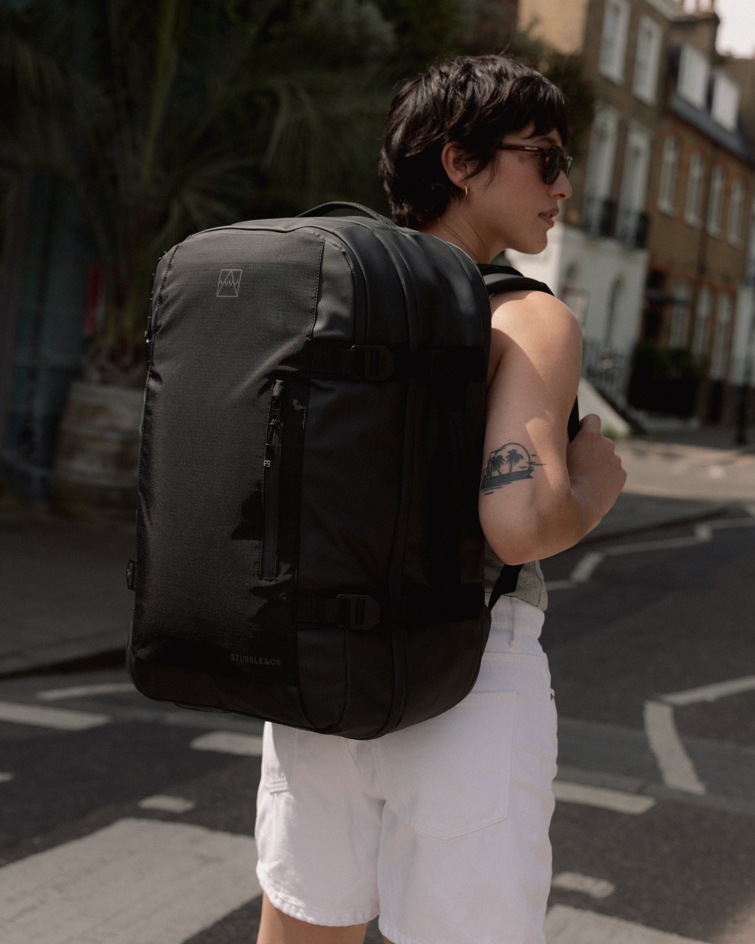 A woman wearing sunglasses and a Travel Backpack in All Black, standing on a sunny street.