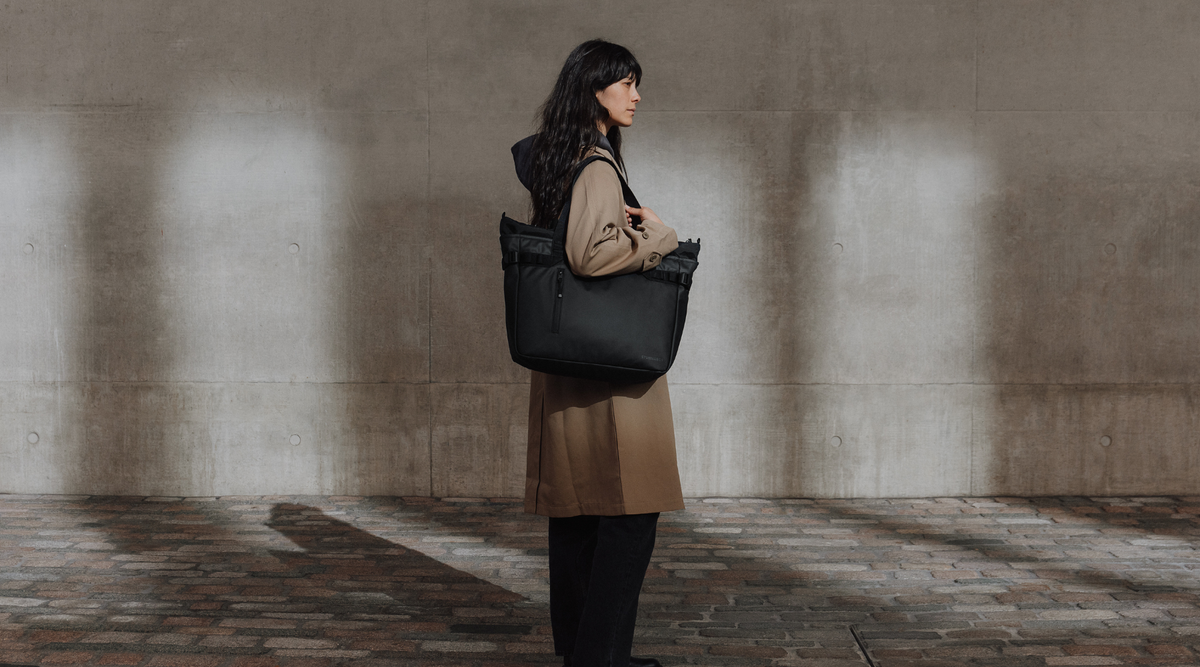 Woman in trench coat carrying the Laptop Tote 20L in All Black against a concrete wall. 