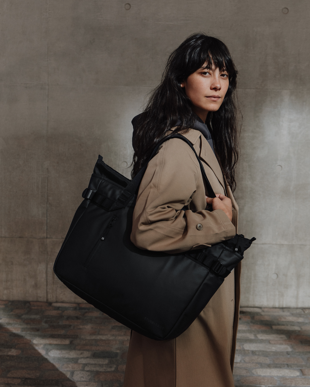 Woman in a beige trench coat carrying the black Laptop Tote against a concrete wall.