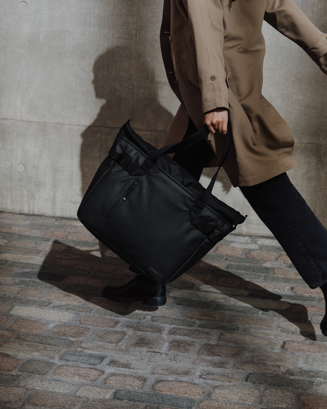 Person in a trench coat carrying the Laptop Tote 20L in All Black while walking.