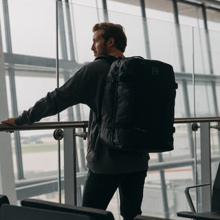 Back view of a man wearing The Adventure Bag i.n All Black while standing in an airport.