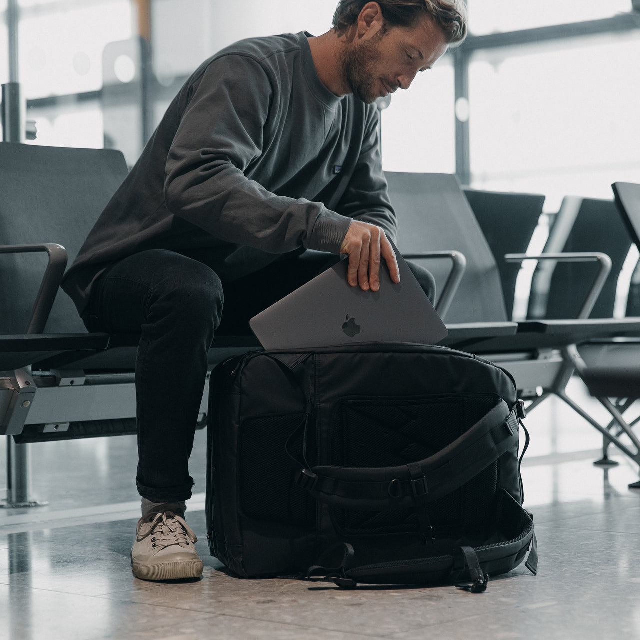 A man sat down in an airport, putting a laptop into The Adventure Bag in All Black