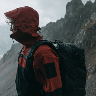 Side view of a man wearing an orange rain coat and The All Black Adventure Bag in the rain, while stood on a mountain side,