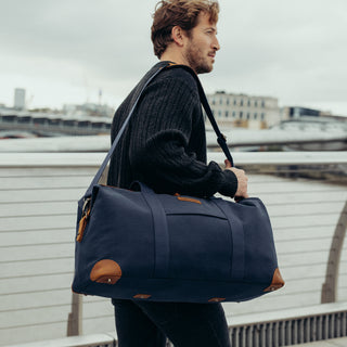 A man walking on a bridge with The Weekender in Navy over his shoulder. 