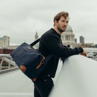 A man from the side leaning on a bridge wall with The Weekender in Navy over his shoulder.