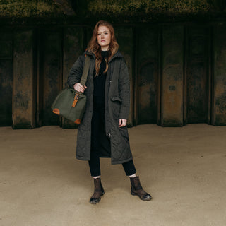 A women stood on a beach wearing a long black coat and The Weekender in Olive over her shoulder.