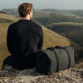 A man with his back to the camera, sat on a rock over looking a hillside, with The Weekender in Pirate next to him.