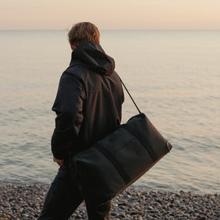 A man wearing a black coat with The Weekender in Pirate over his shoulder, looking out to sea on a pebbled beach.