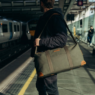 A man stood on a train platform with The Weekender in Olive over their shoulder.