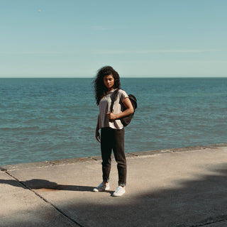 A women facing the camera wearing a grey top and the All Black Backpack Mini over one shoulder. Stood in front of the sea.