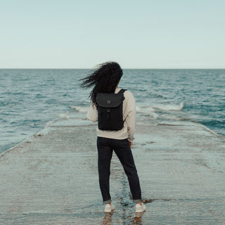 A women with her pack to the camera while standing on a slipway into the sea. Sh e is wearing The Backpack Mini in All Black.