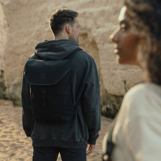 A man in the background with his back to the camera wearing The Backpack Mini in All Black while standing on a beach. In the foreground a women is just walking into shot.